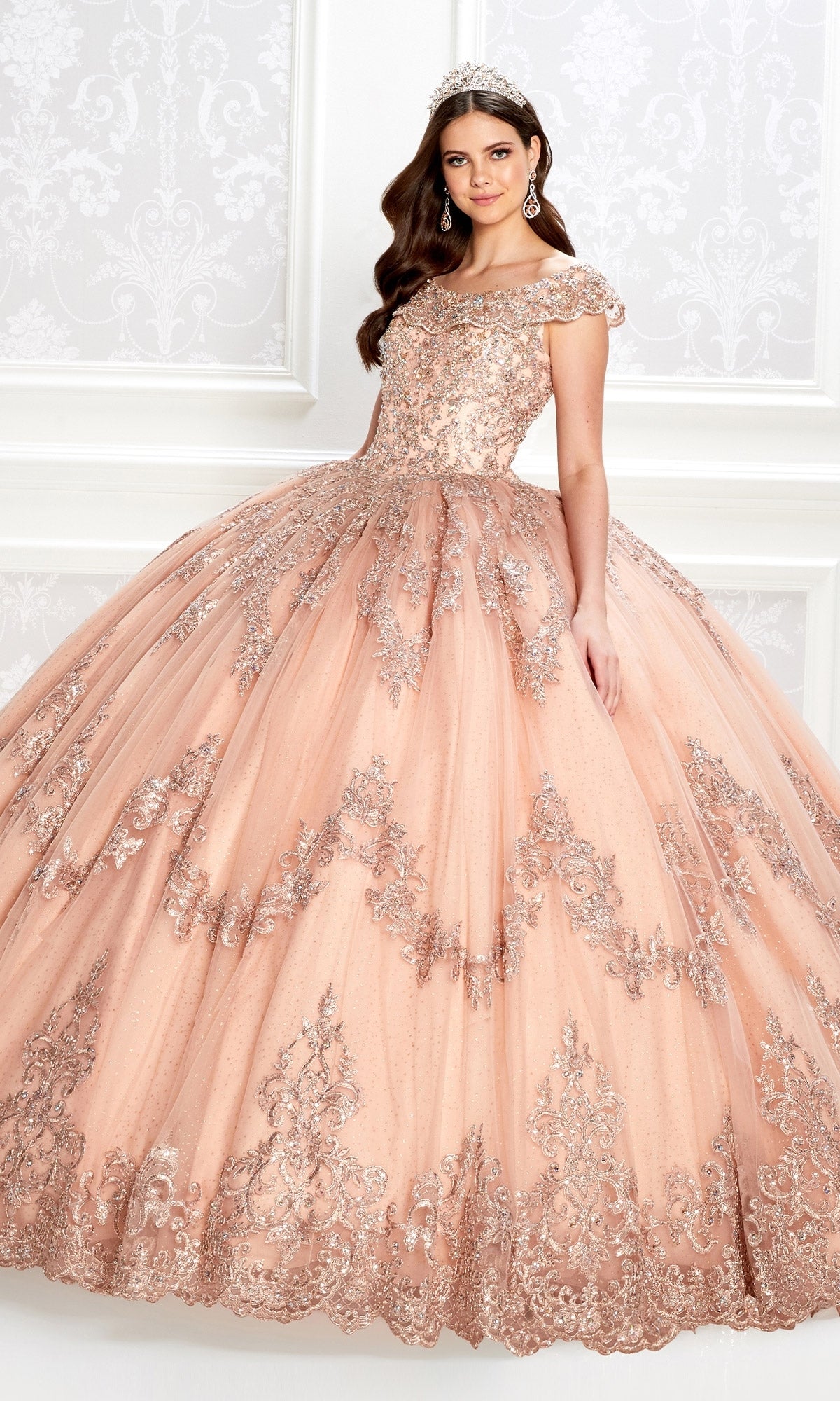A woman wearing the Quinceanera Dress PR22035 By Princesa, featuring a peach hue, corset bodice, silver embroidery, and full skirt, is accessorized with a tiara and earrings as she stands before an ornate white wall.